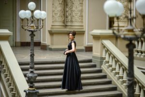 Young woman in an elegant black gown standing alone on a grand staircase, confidently posing for a prom alone celebration.
