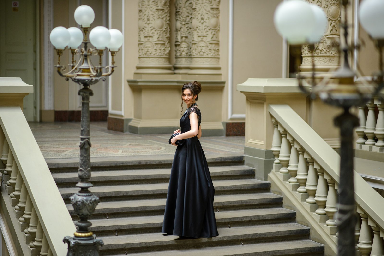 Young woman in an elegant black gown standing alone on a grand staircase, confidently posing for a prom alone celebration.